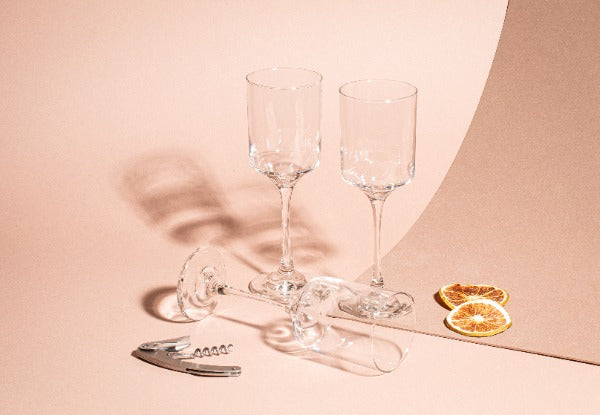 Tres copas de vidrio para vino sobre fondo beige, dos en posición vertical y una acostada, acompañadas de un sacacorchos metálico y dos rodajas de naranja deshidratada. Colección Río