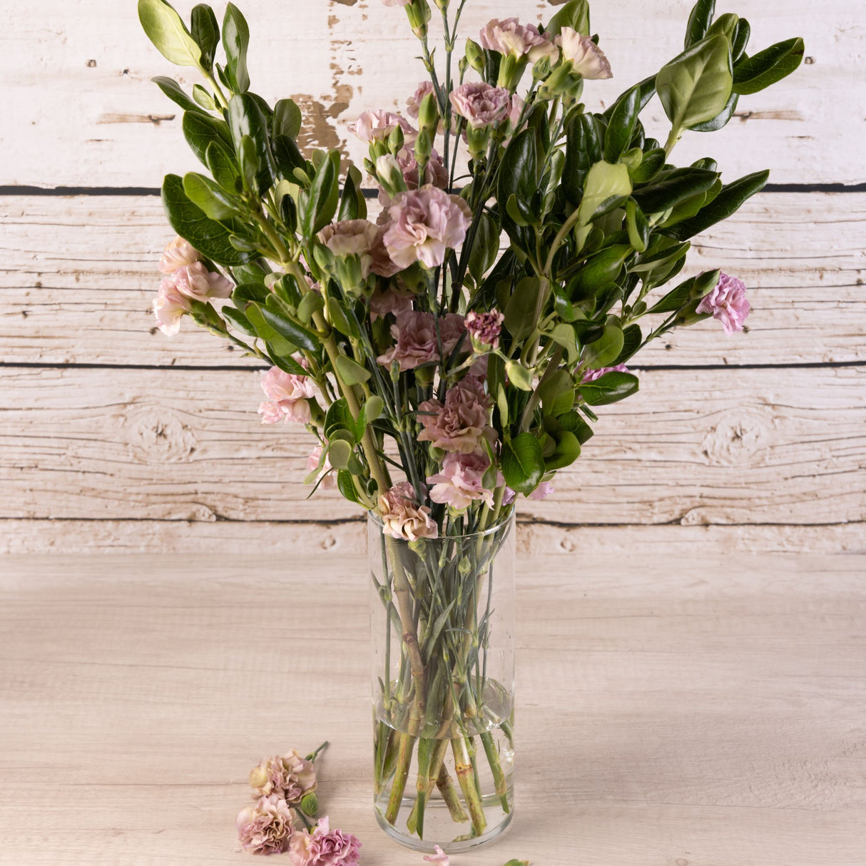 Florero de vidrio transparente con forma cilíndrica, con ramo de flores rosadas sobre superficie y fondo de madera blanca rústica.