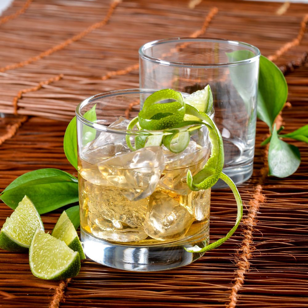 Dos vasos rocks de vidrio transparente – uno con cóctel dorado, hielo y espiral de limón, otro vacío, sobre superficie tejida con hojas verdes y limones partidos