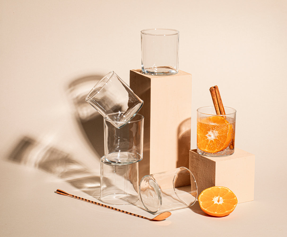 Vasos Ríos Rocks dispuestos sobre bloques beige, acompañados de una bebida con rodaja de naranja y canela, y una mitad de mandarina al frente.