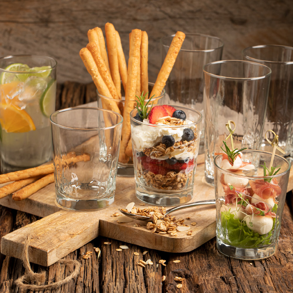Siete vasos de vidrio, para bebidas y jugos de diferente tamaño, puestos sobre bandeja de madera y decorados con alimentos
