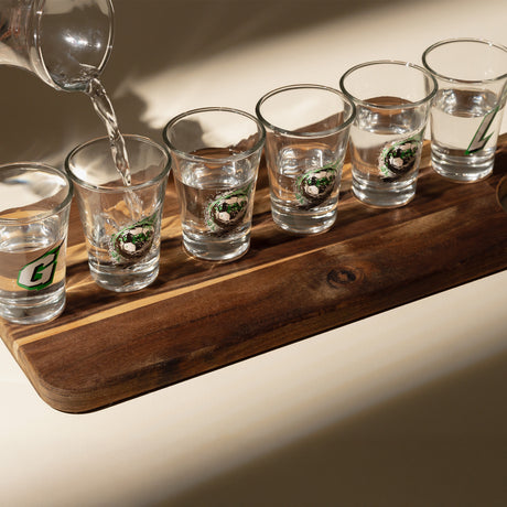 Vasos shot alineados en tabla de madera con jarra vertiendo agua en ambiente iluminado