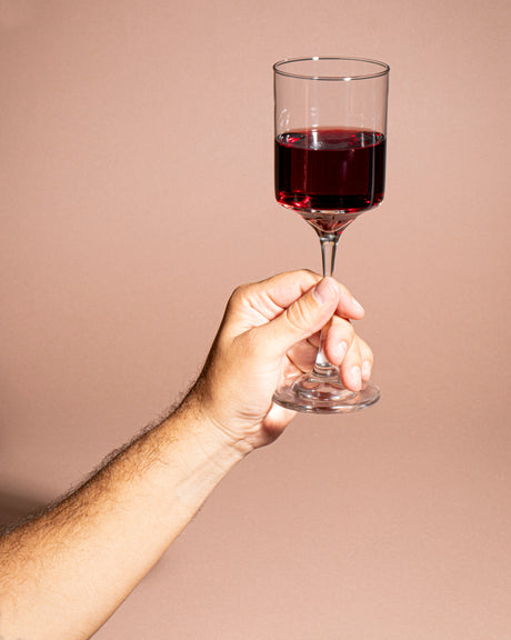 Mano sosteniendo una copa de vidrio transparente llena de vino tinto sobre un fondo beige.  Colección Río