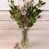 Florero de vidrio transparente con forma cilíndrica, con ramo de flores rosadas  sobre superficie y fondo de madera blanca rústica.