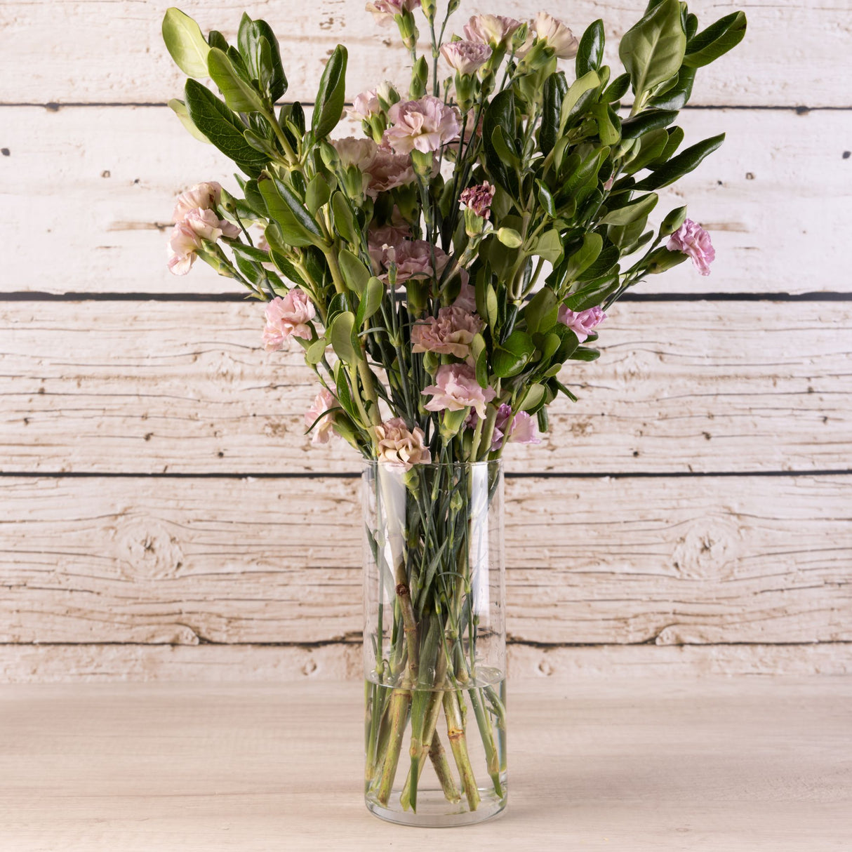 Florero cilíndrico de vidrio transparente con ramo de flores rosadas y follaje verde, sobre superficie de madera blanca rústica