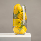 Un florero conico de vidrio transparente con limones amarillos y ramas de romero sumergidos en agua, sobre superficie blanca y fondo gris.