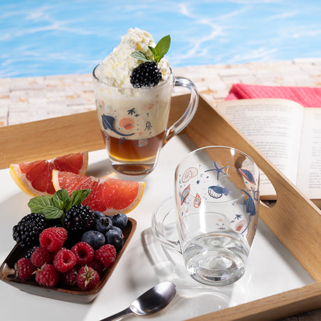 Dos mugs de vidrio transparente con diseño de corales sobre bandeja con frutas
