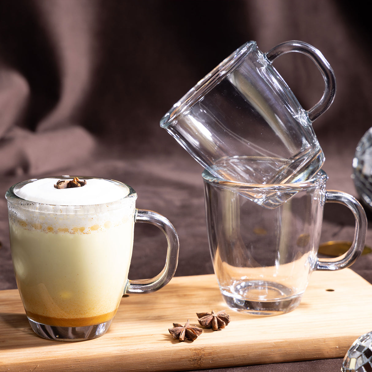 Mug de vidrio London con bebida tipo latte con espuma y anís en mesa, junto a tazas apiladas