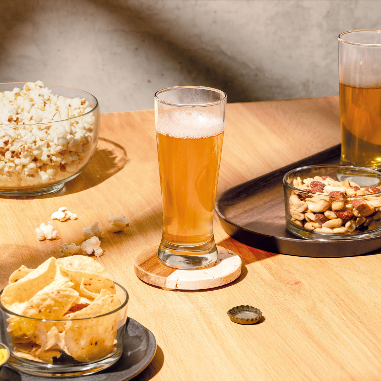 Vaso de vidrio alto con cerveza dorada, snacks en bowls sobre superficie de madera