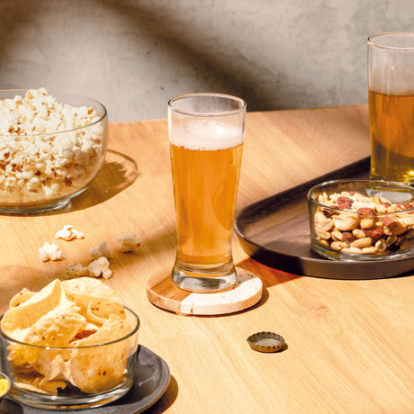 Vaso de vidrio alto con cerveza dorada acompañado de snacks en bowls sobre superficie de madera en ambiente casual