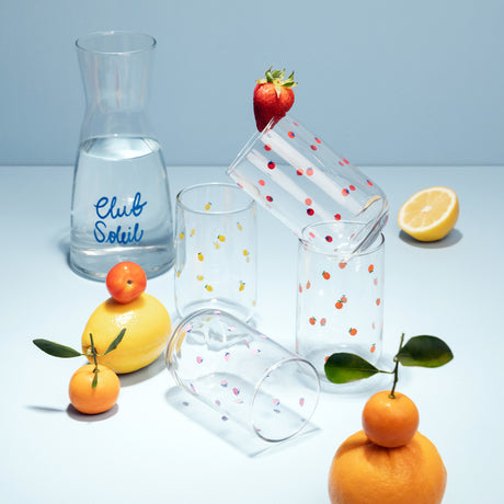 Un decanter con letras Club solei. 4 vasos de vidrio con figuras de frutas.
