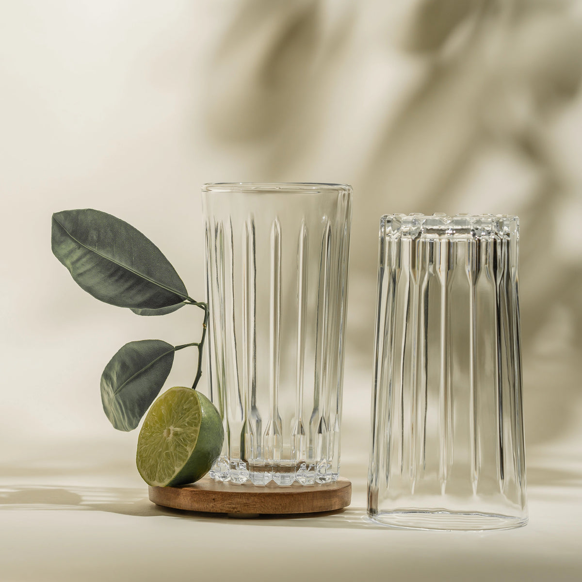 Dos vasos altos de vidrio Bali largos sobre fondo beige claro, uno apoyado en un posavasos de corcho junto a una rama con hojas y una rodaja de lima, con sombras de hojas proyectadas en la pared.