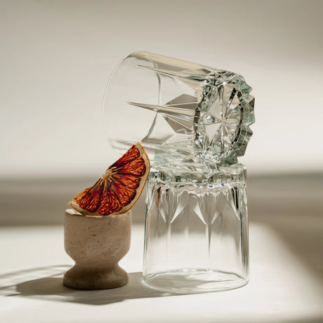 Vaso de vidrio Creta Rocks transparente con textura, acompañado de otro vaso invertido y una rodaja de naranja sanguina sobre superficie clara.