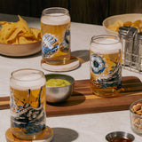Tres vasos de vidrio Lata edición Fútbol con cerveza, sobre mesa con tabla de madera, snacks, nachos y salsas en ambiente de reunión.