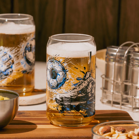 Vaso de vidrio con diseño de balón de fútbol lleno de cerveza, sobre tabla de madera con snacks