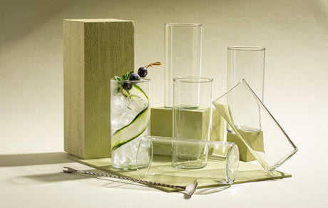 Arreglo artístico de varios vasos de vidrio Rio bebidas colocados sobre una superficie beige, acompañado de un vaso con hielo, pepino en espiral y arándanos, junto a un removedor metálico.