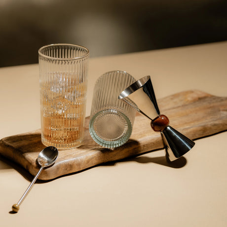 Dos vasos de vidrio Santorini formato largo o para bebidas, uno de pie y otro inclinado, sobre superficie de madera.