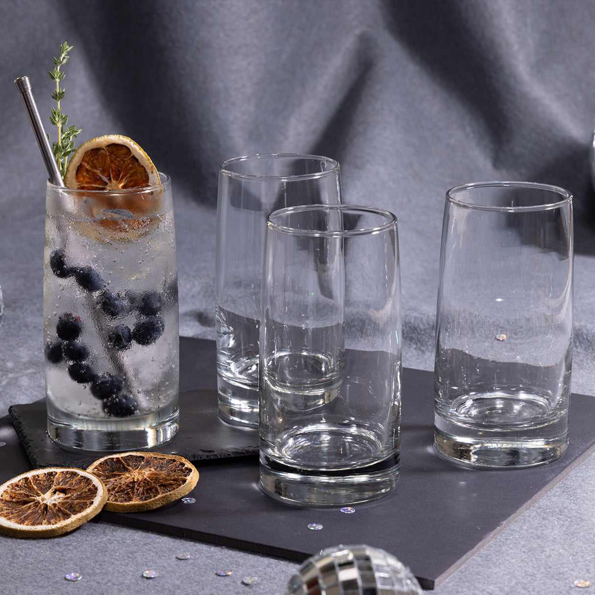 Cuatro vasos de vidrio Toledo sobre una superficie oscura, uno con bebida fría decorada con arándanos, rodaja de naranja deshidratada y hierbas, junto a otros vasos vacíos.