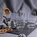 Cuatro vasos de vidrio Toledo sobre una superficie oscura, uno con bebida fría decorada con arándanos, rodaja de naranja deshidratada y hierbas, junto a otros vasos vacíos.