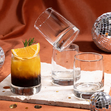 Tres vasos de vidrio Toledo sobre una superficie clara, uno con cóctel de capas y rodaja de naranja, y tres vasos vacíos, con bolas disco decorativas alrededor.